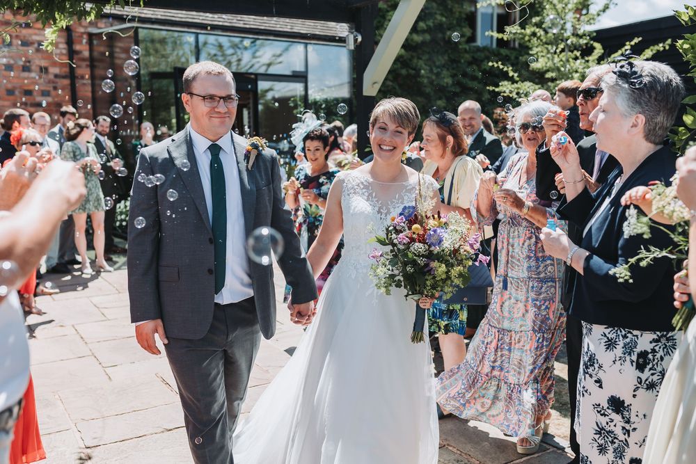 Wedding guests line a sidewalk outside while blowing bubbles as a couple walks through during their celebration.