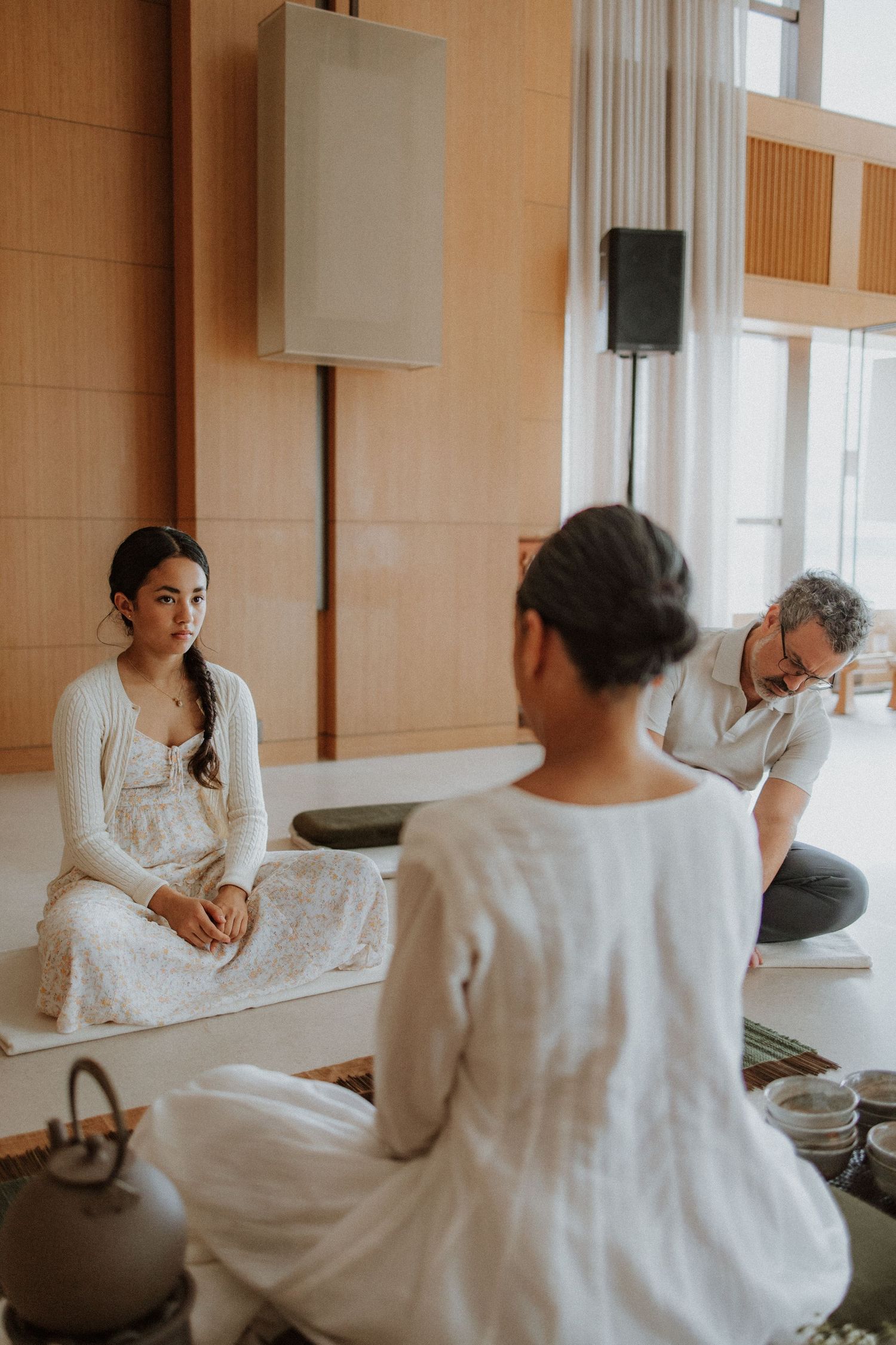 People wearing white dresses sit together in a minimalist room with wood paneled walls and speakers mounted above.