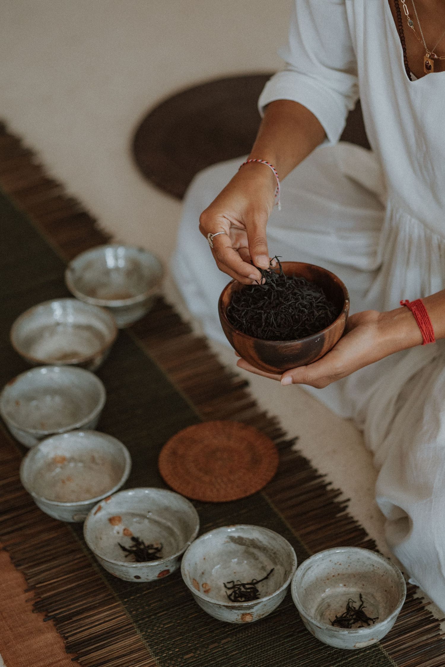 Someone in white clothing holding a bowl of loose tea leaves with small ceramic bowls arranged for tea tasting on a bamboo mat.