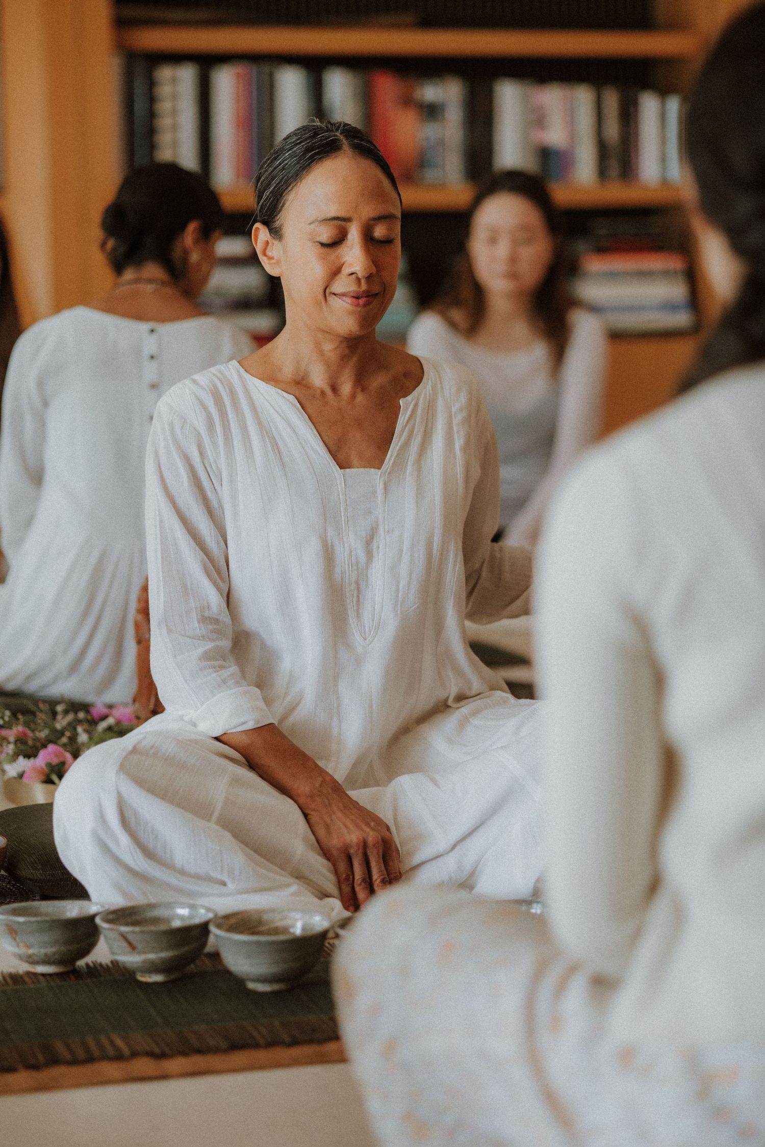 A person in white clothing sits peacefully in meditation pose during a traditional tea ceremony gathering.