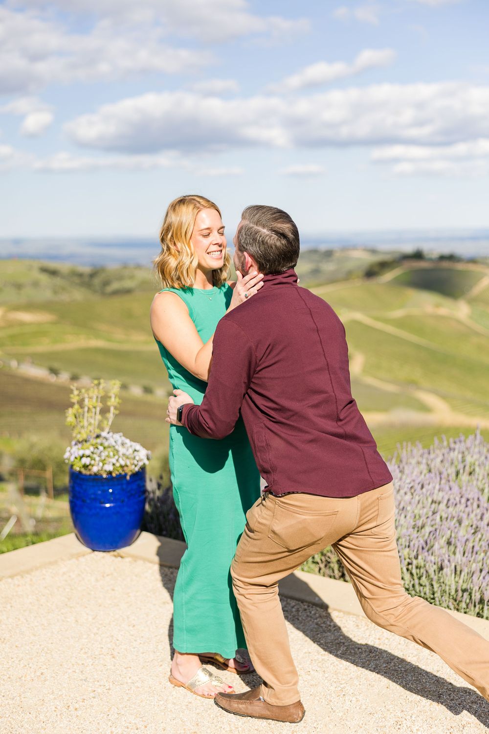 A couple shares a romantic moment on a hilltop overlooking rolling green hills and vineyards with a blue flower pot nearby.