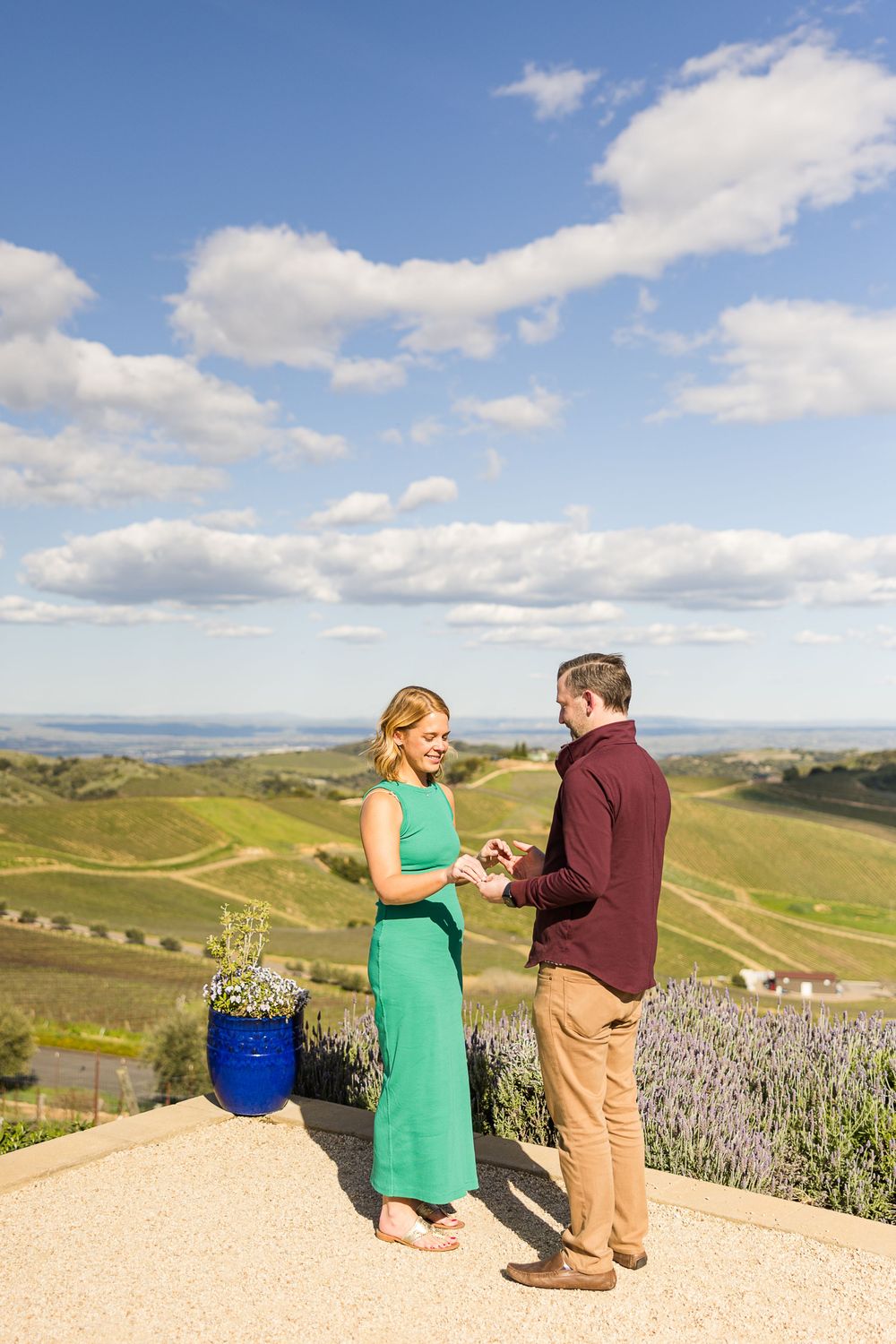 Two people standing on a hilltop overlook with rolling green hills and blue skies with puffy white clouds in the background.
