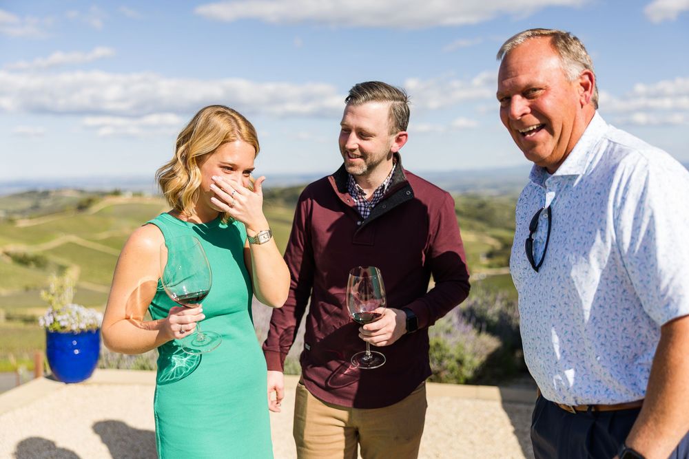 Three people enjoying wine and laughs together on a scenic hilltop with rolling landscapes in the background.