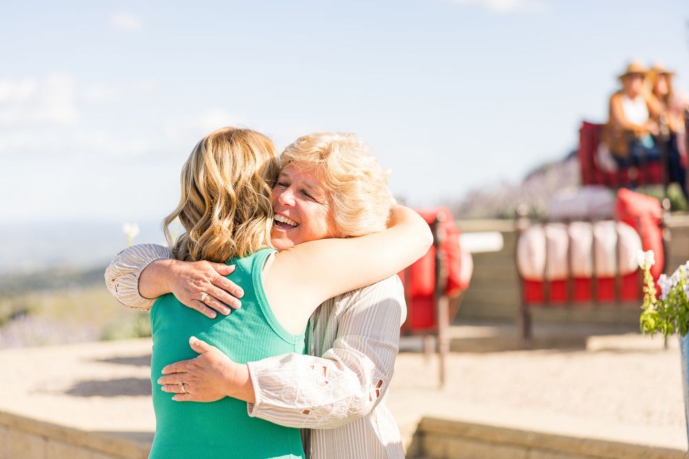 Two people embrace in a warm hug outdoors with mountains and bright blue sky in the background.