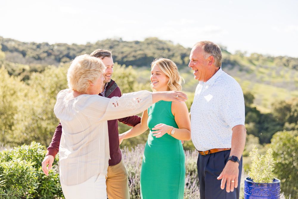 A group of people laugh together while standing outdoors in a sunny hillside setting with green foliage.