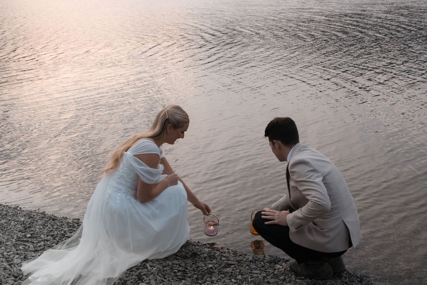 A couple crouches by the water's edge at sunset in formal wedding attire, creating a serene lakeside moment.