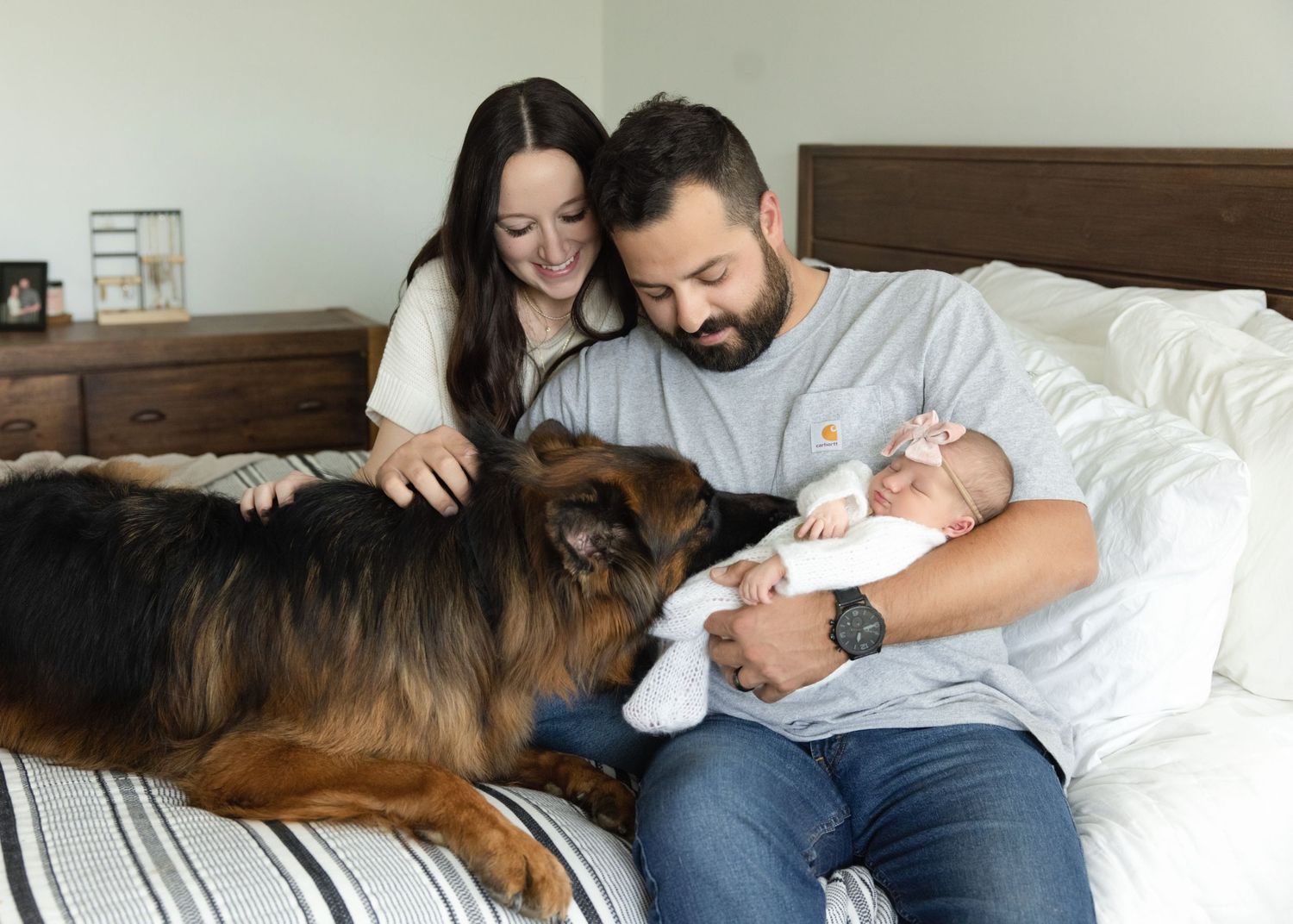 A large brown dog lies on a bed with a couple as they bond and spend quality time together with their newborn baby girl in a bedroom.
