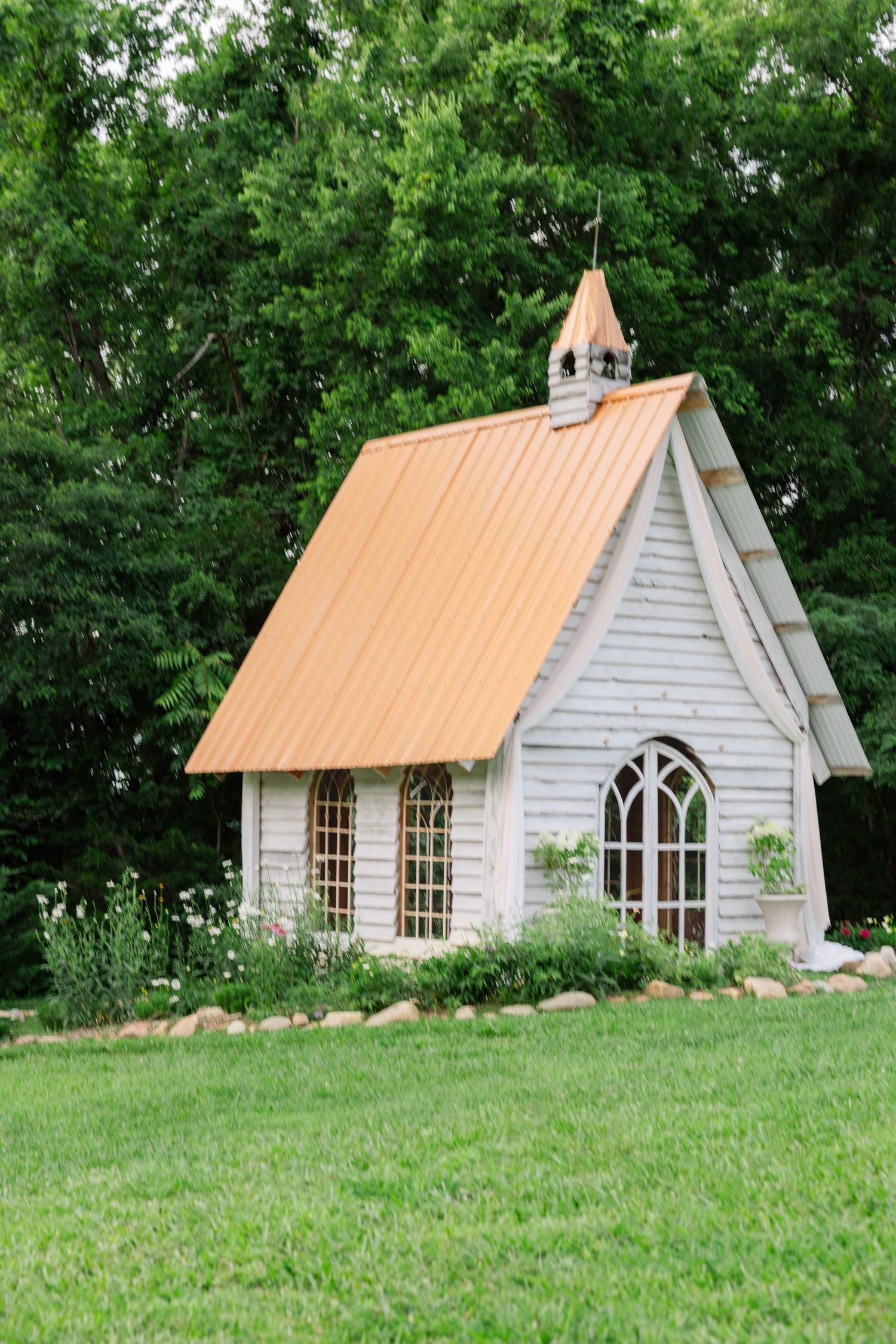 Charming white country chapel with copper-colored metal roof and gothic windows sits nestled among green trees and landscaping.