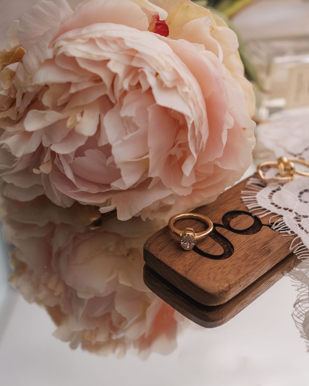Wedding rings rest on a wooden box beside soft pink peonies and lace details.
