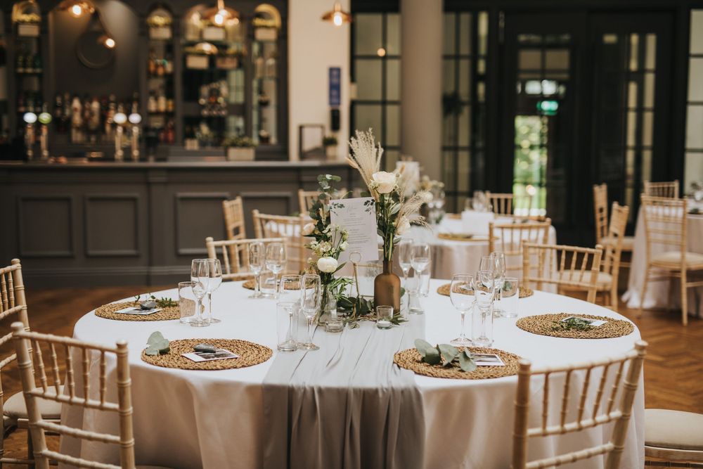 Elegant wedding reception tables with white linens, gold chargers, and chiavari chairs in a bright dining room.