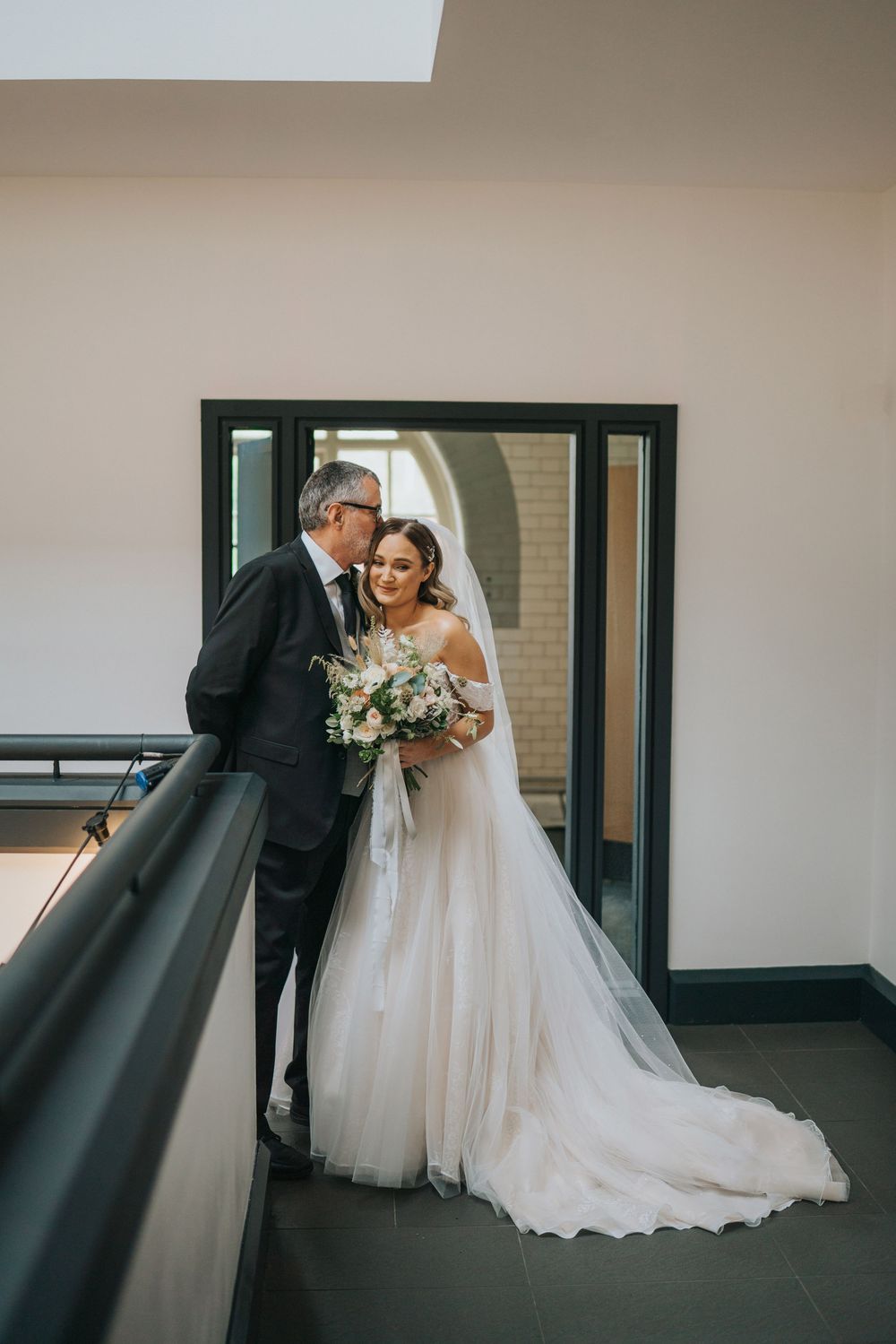 Two people share an intimate moment in a modern hallway, with a flowing white wedding dress and formal attire.