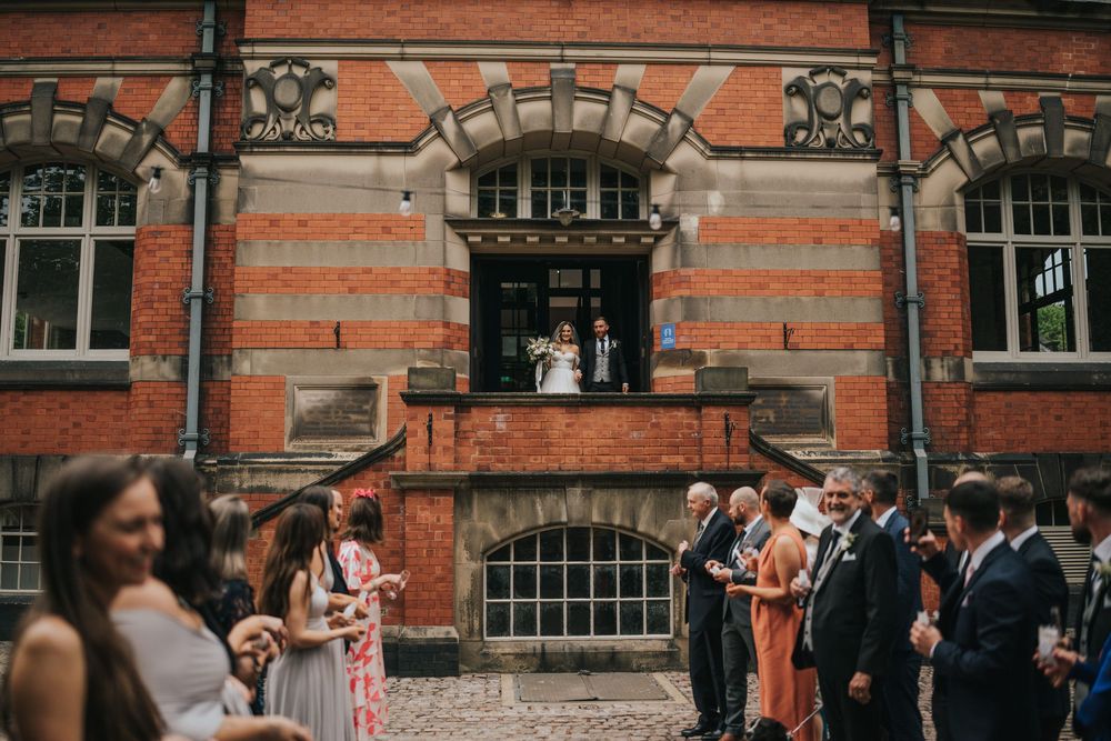 People gather outside a historic red brick Victorian building with ornate stone archways and window details.