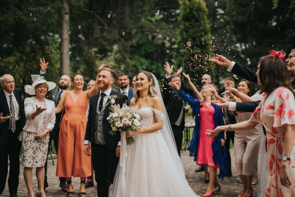 Wedding guests celebrate and throw rose petals at a joyful newlywed couple during outdoor ceremony exit.