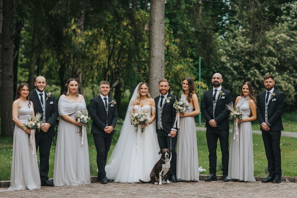 Wedding party in formal attire stands outdoors with a black and white dog on paved path surrounded by tall trees.