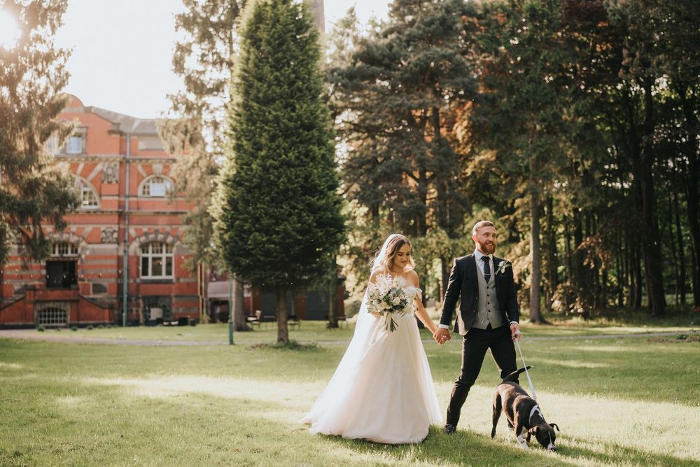 A couple in wedding attire stands on a green lawn near a brick building with their small dog on a fall day.