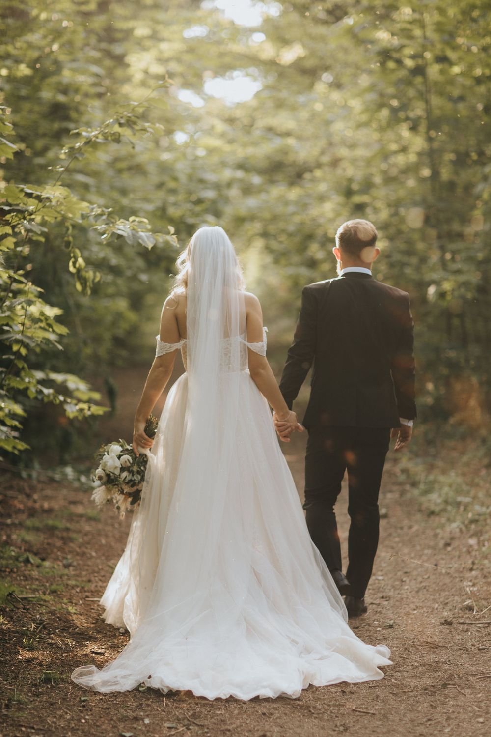 Bride and groom walk hand in hand down a sunlit forest path, her flowing white gown trailing behind them on the dirt trail.