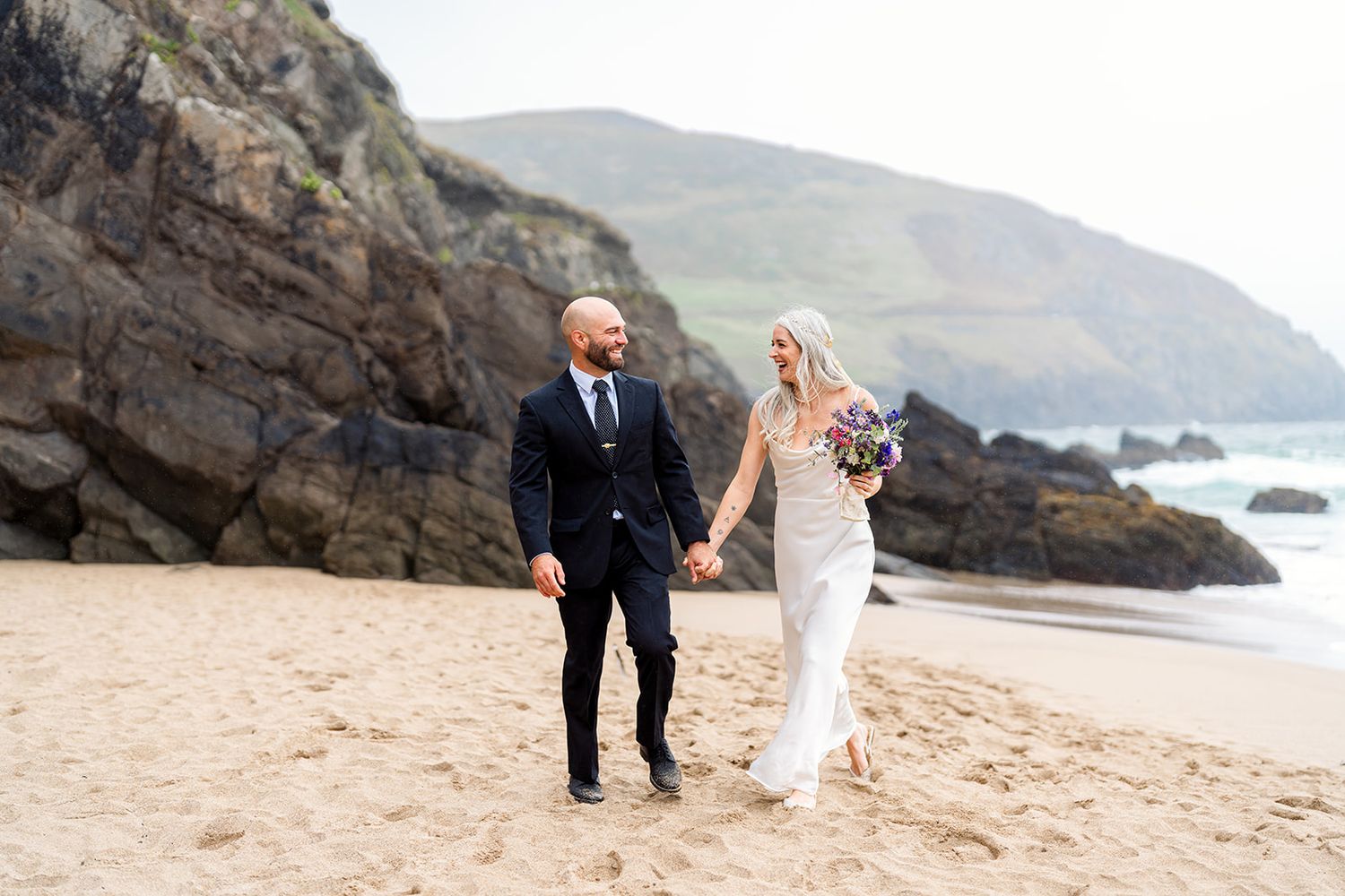A couple in wedding attire walk hand in hand on a sandy beach with rocky cliffs and coastline in the background.