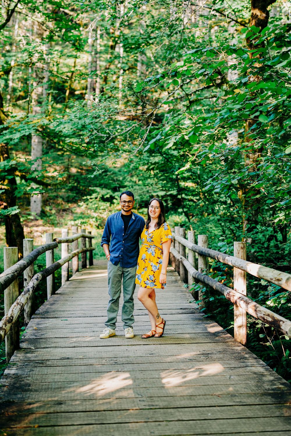 Couple standing together on a wooden boardwalk trail surrounded by lush green forest on a sunny day.