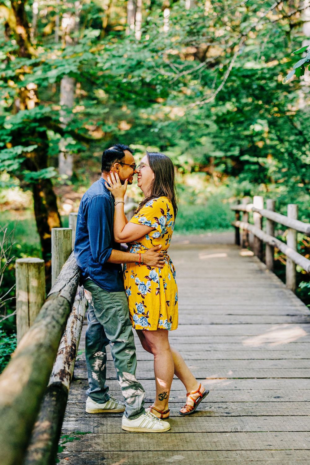 A couple shares a romantic moment on a wooden footbridge surrounded by lush green forest scenery.