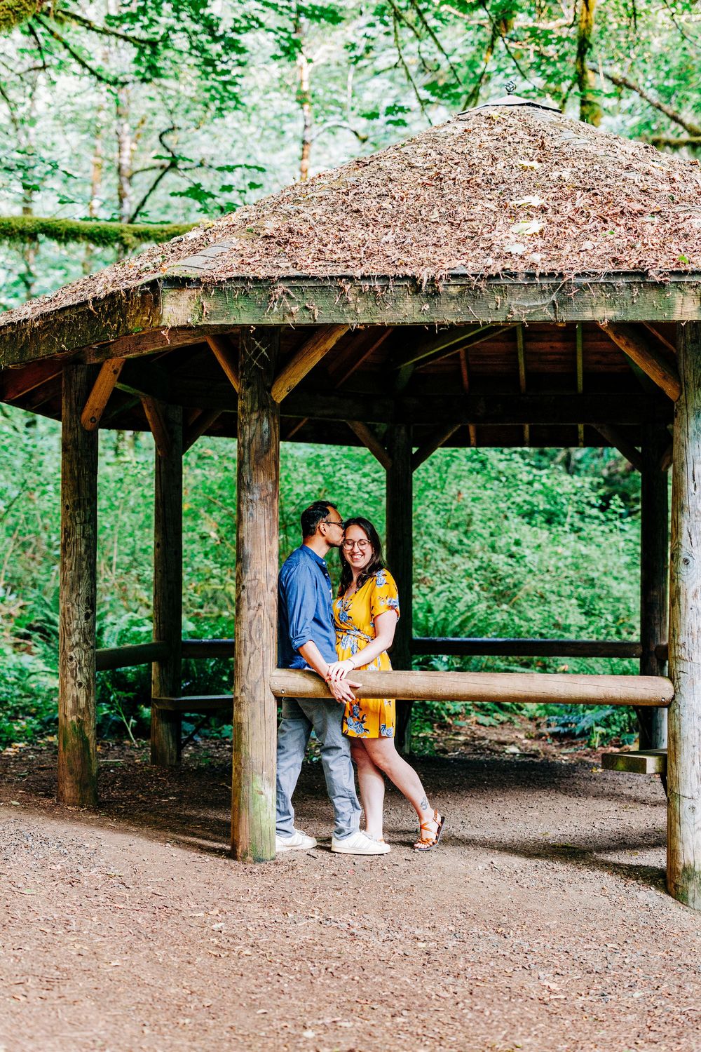 A couple embraces under a rustic wooden gazebo with a thatched roof surrounded by lush green forest.