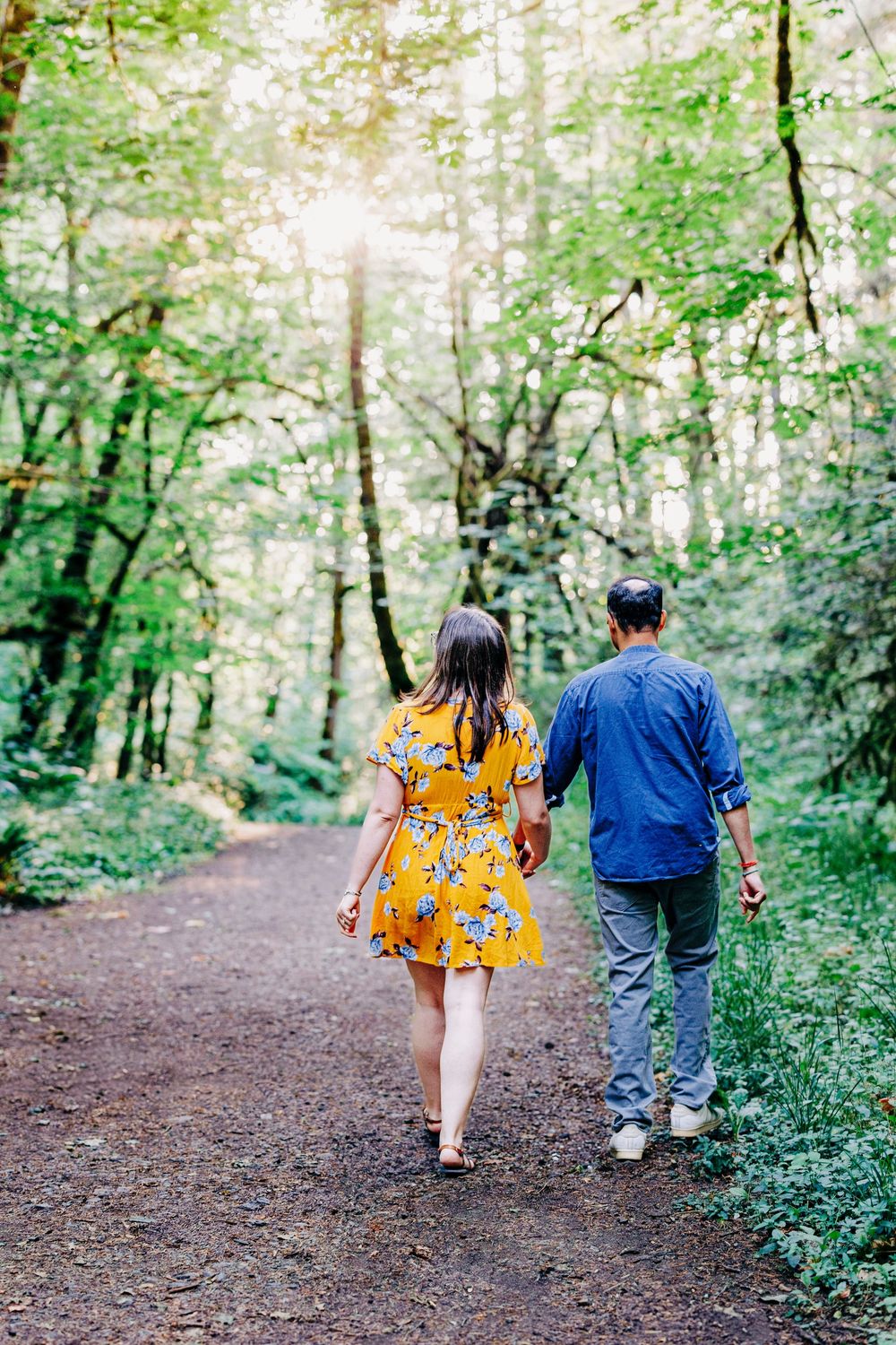 A couple walks together on a forest trail surrounded by lush green trees and dappled sunlight.