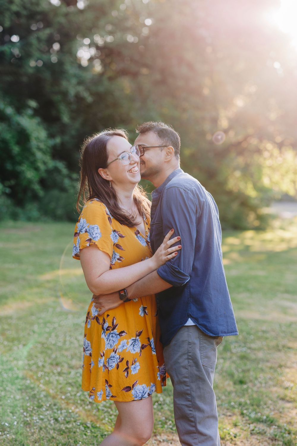 A couple embraces in a sunlit garden, with the woman wearing a yellow floral dress and the man in a navy shirt.