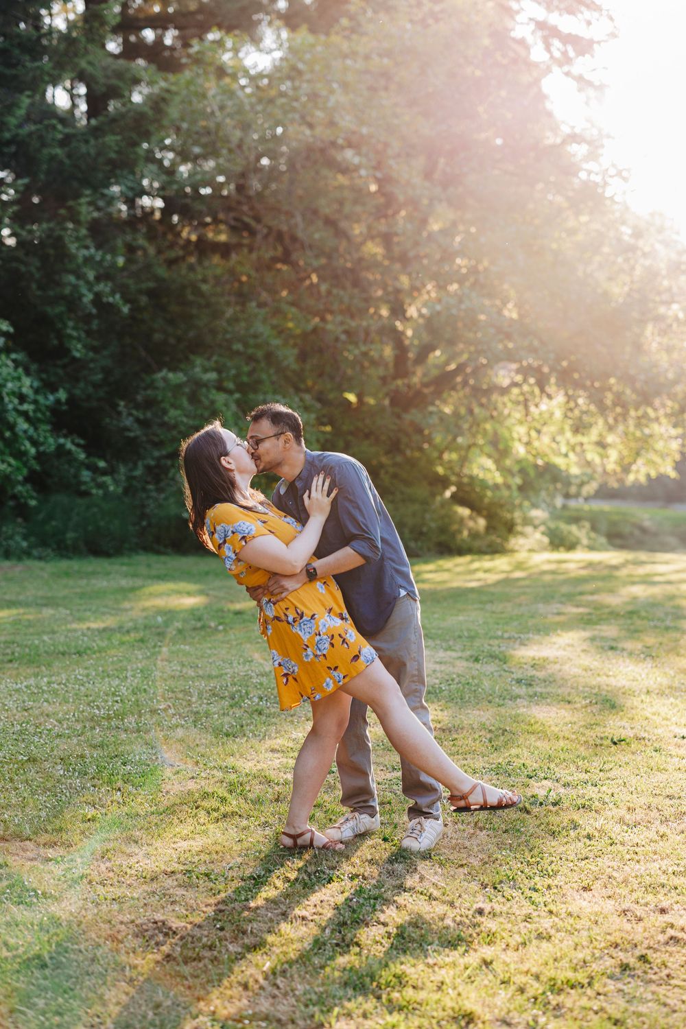 A couple embraces in a romantic dip pose as golden sunlight streams through trees in a backyard garden setting.