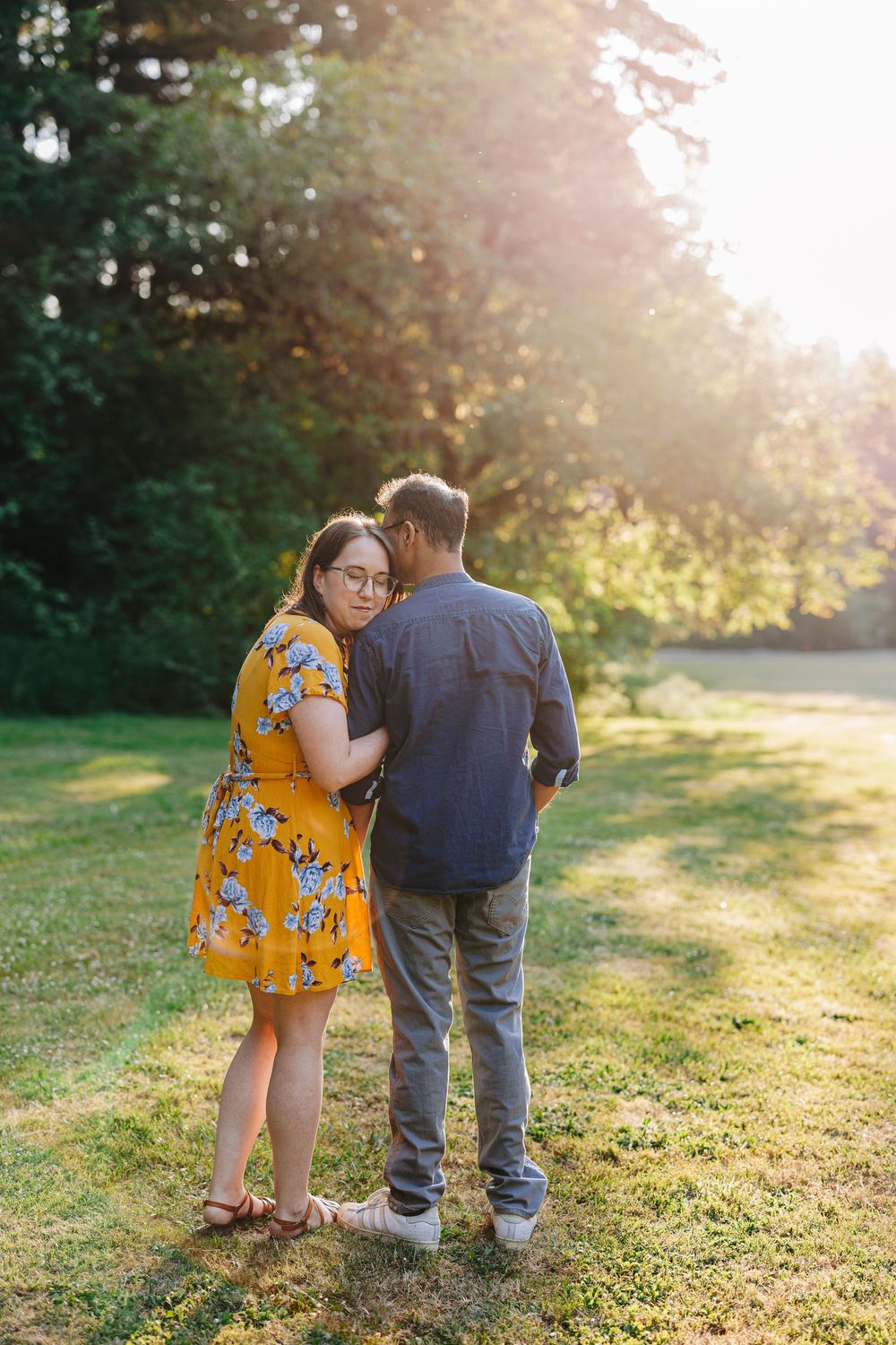 Romantic couple embracing on grassy lawn at sunset with trees and golden sunlight in background.