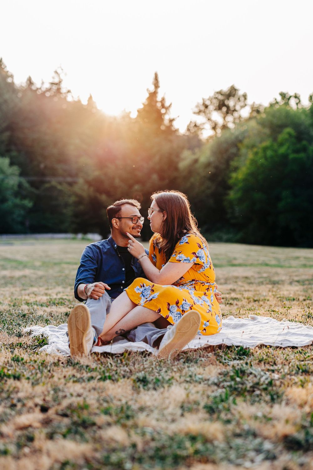 A romantic couple shares an intimate moment on a picnic blanket at sunset in a grassy field.