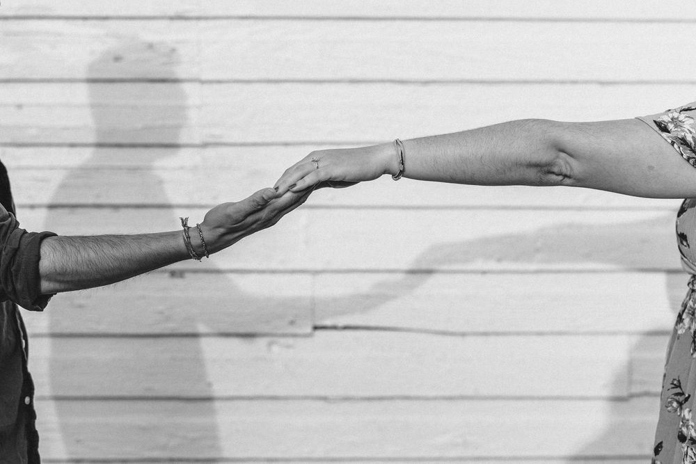 Two arms reach out from opposite sides to hold hands against a white wooden wall background.