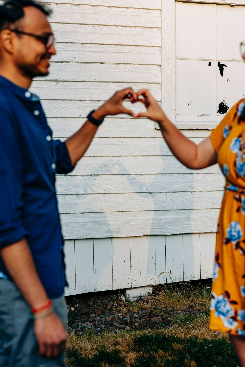 Two people in blue and yellow outfits making a heart shape with their hands against a white wall.