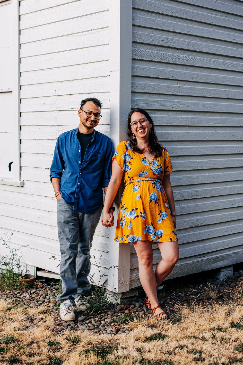 A couple stands together against a white wooden wall, wearing a blue shirt and yellow floral dress.