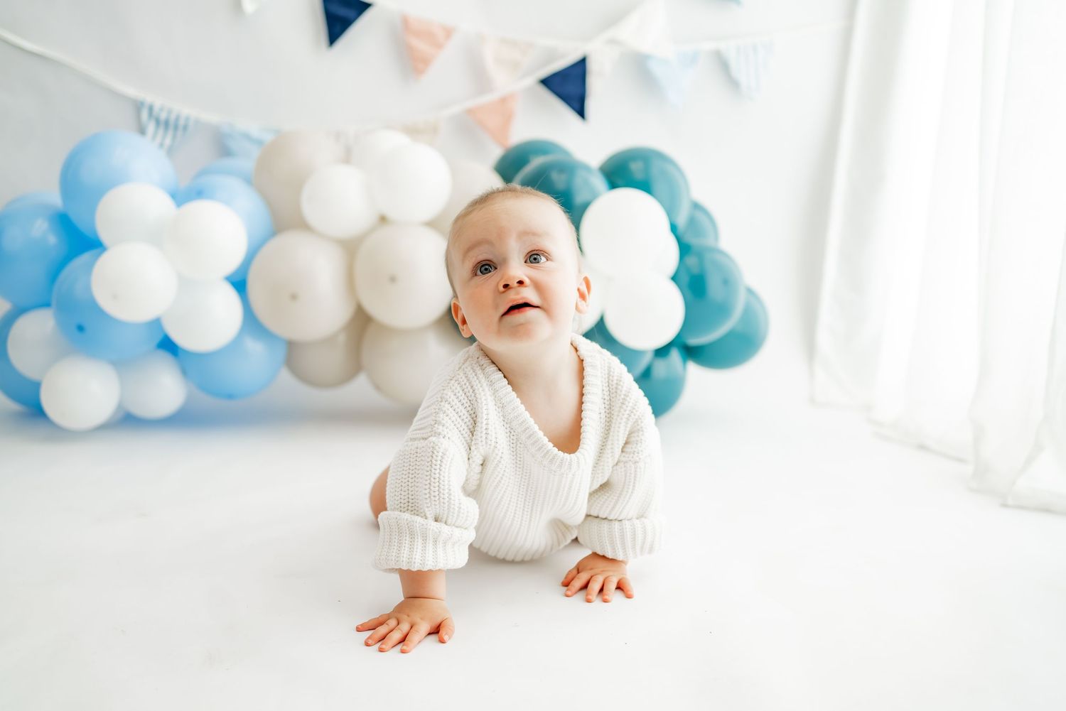 Baby in white outfit crawls on floor with blue and white balloon decorations in bright modern room.