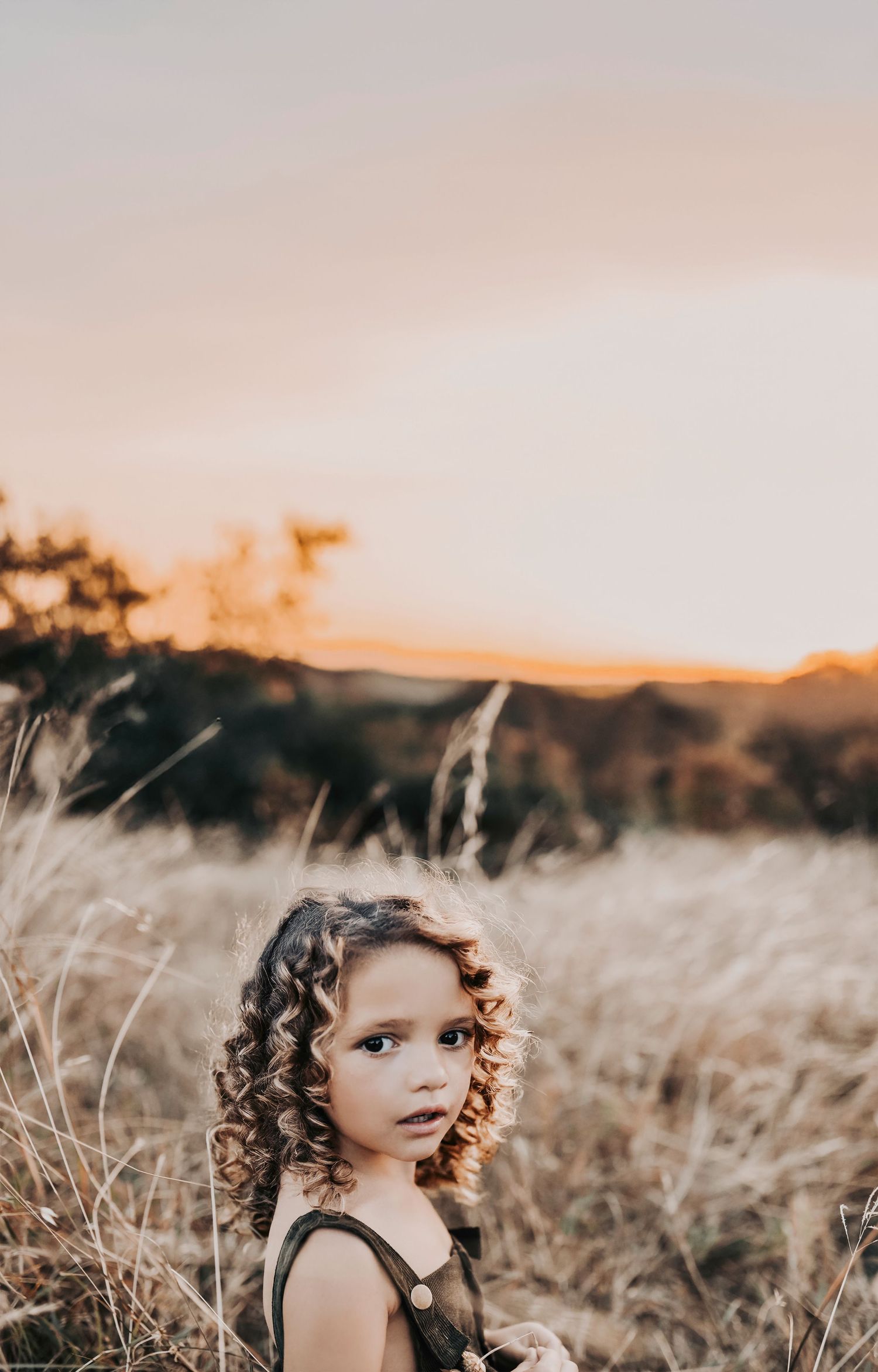 a curly haired little girl stands in a field of golden grass at sunset for her family photos