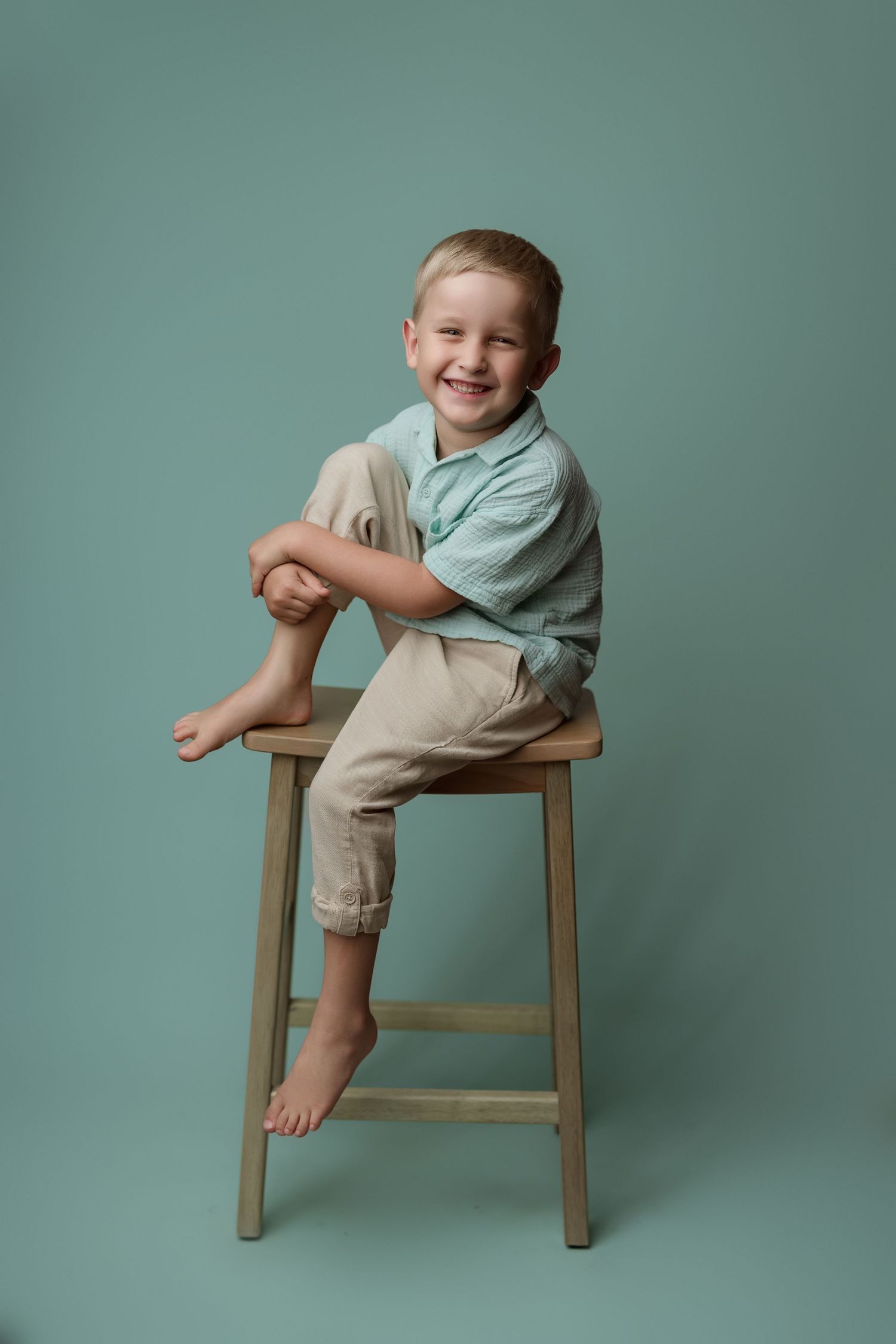 A playful child wearing a mint green shirt and khaki pants sits and laughs while posed on a brown wooden chair.