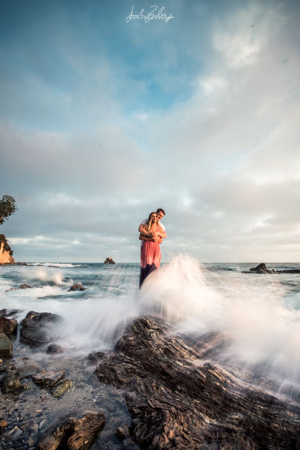 Dramatic coastal portrait with waves crashing against rocks as couple embraces under cloudy sky at the little corona del mar tide pools.