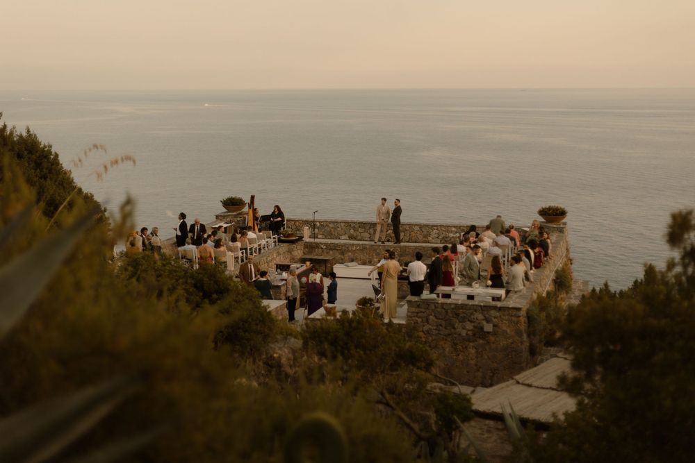 Visitors gather at a scenic overlook on the coast during sunset with multiple boats visible in the ocean below.