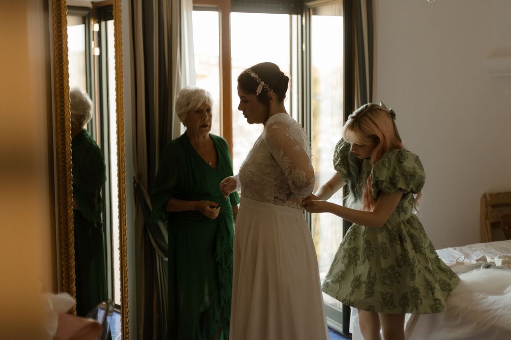 Three people gather in a sunlit room, two wearing elegant gowns in white and green while preparing for a special occasion.