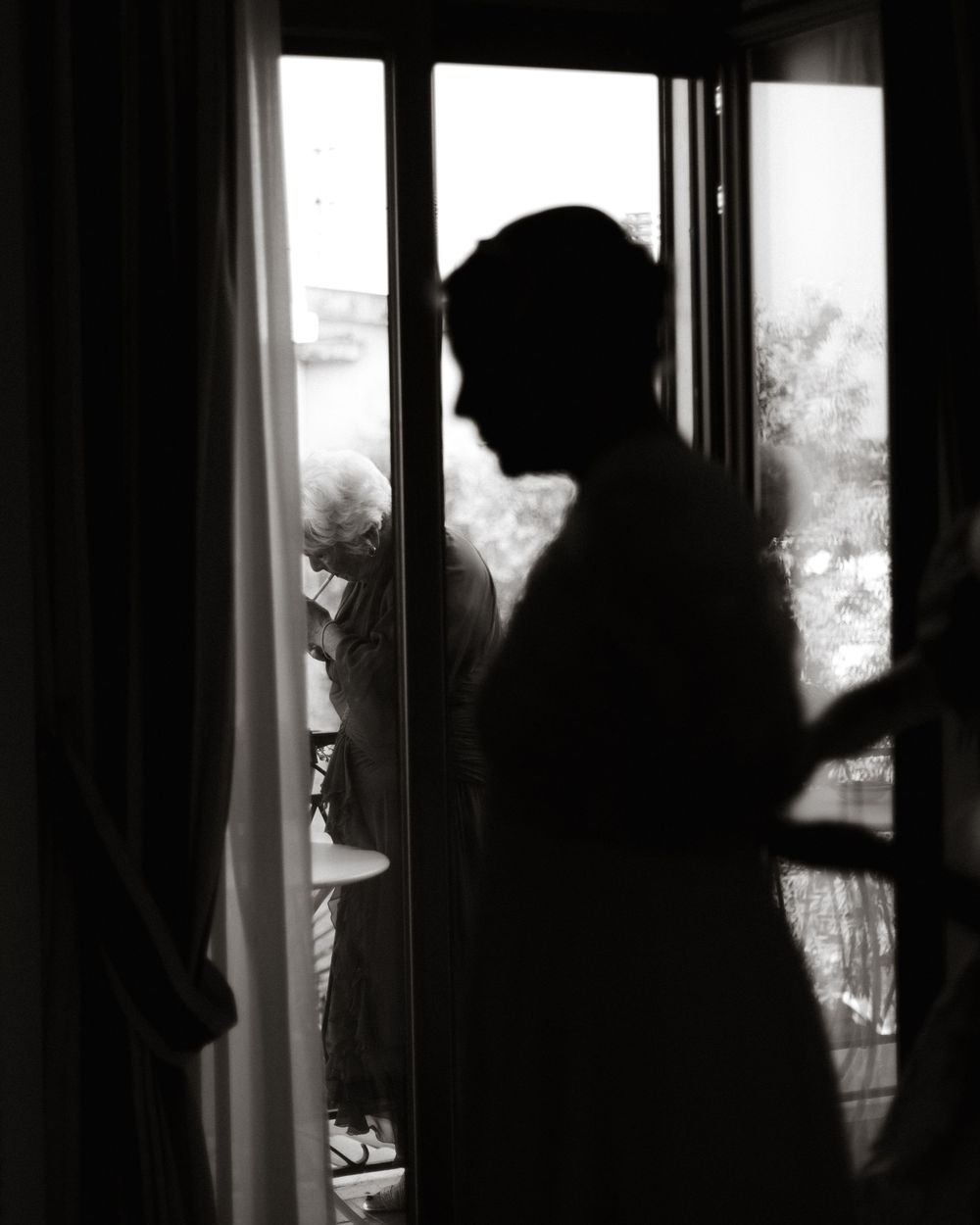 A silhouetted figure stands in a dark interior looking through curtained windows, creating a dramatic contrast of light and shadow.