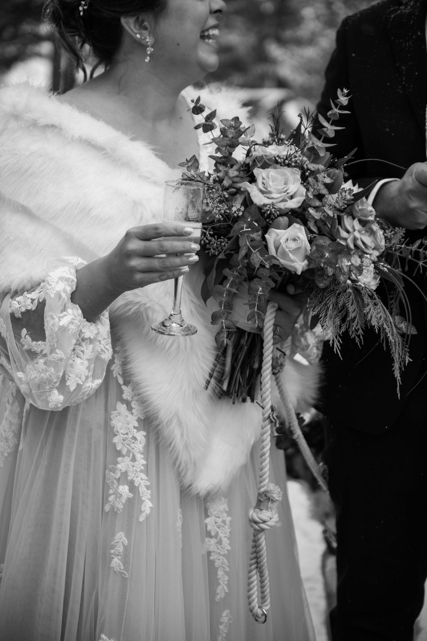 Black and white wedding photo showing a bridal bouquet with roses and a champagne glass against a white lace dress.