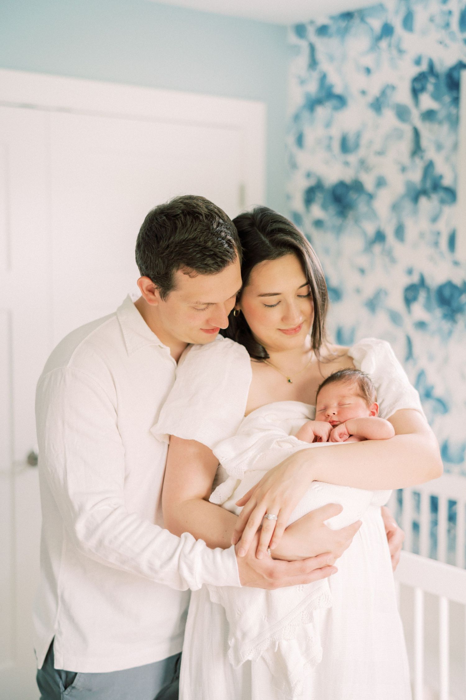 A couple in white clothing stands together in a room with blue floral wallpaper while holding a newborn baby.
