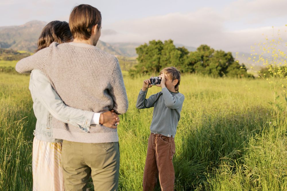 Hands-on family photo session. Family stand together in a sunlit meadow with mountains and trees in the background on a warm afternoon.
