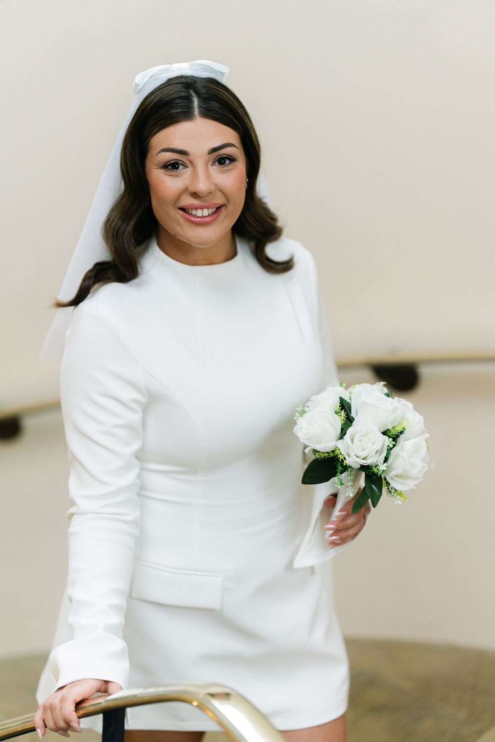 A bride in a sleek white long-sleeved dress holds a white peony bouquet against a neutral background.