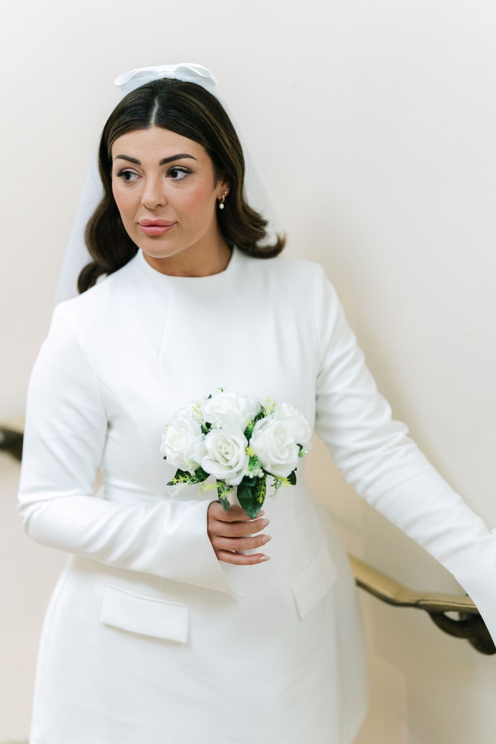 Modern bride in elegant white long-sleeved dress holding simple white rose bouquet against light background.
