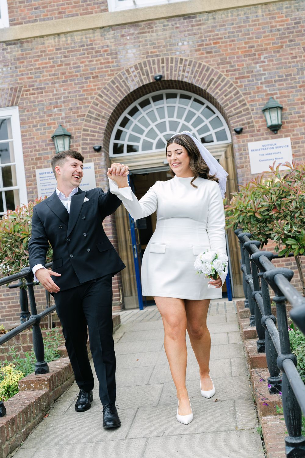 Bride in short white dress and groom in black suit celebrate joyfully while walking down church steps on wedding day.