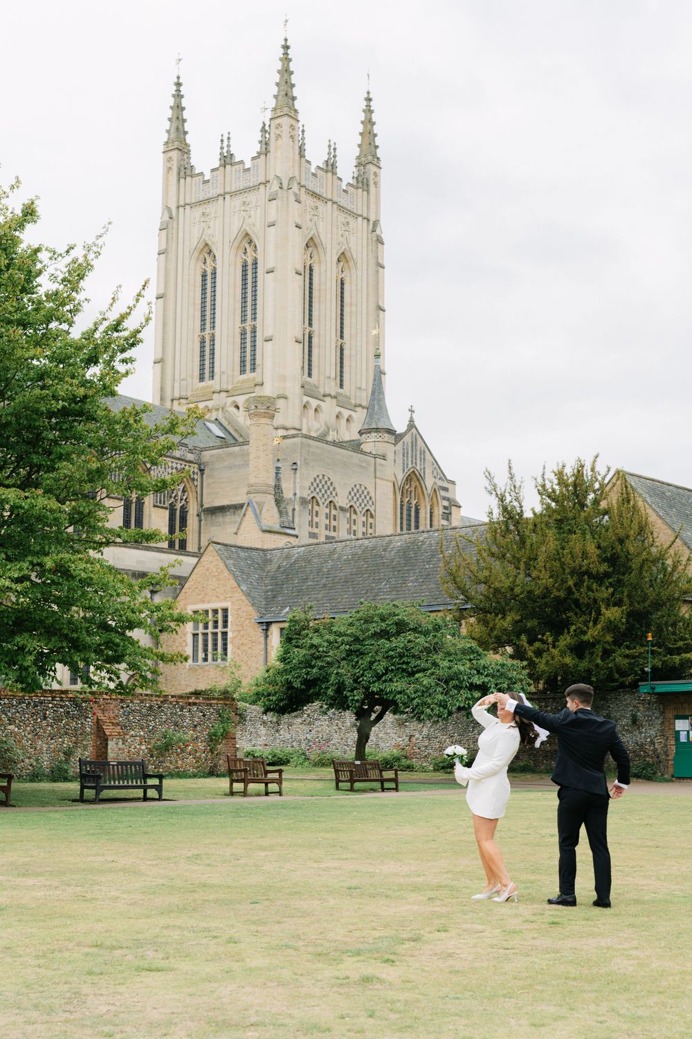 Two figures embrace on a lush green lawn with a grand Gothic cathedral spire towering in the background.