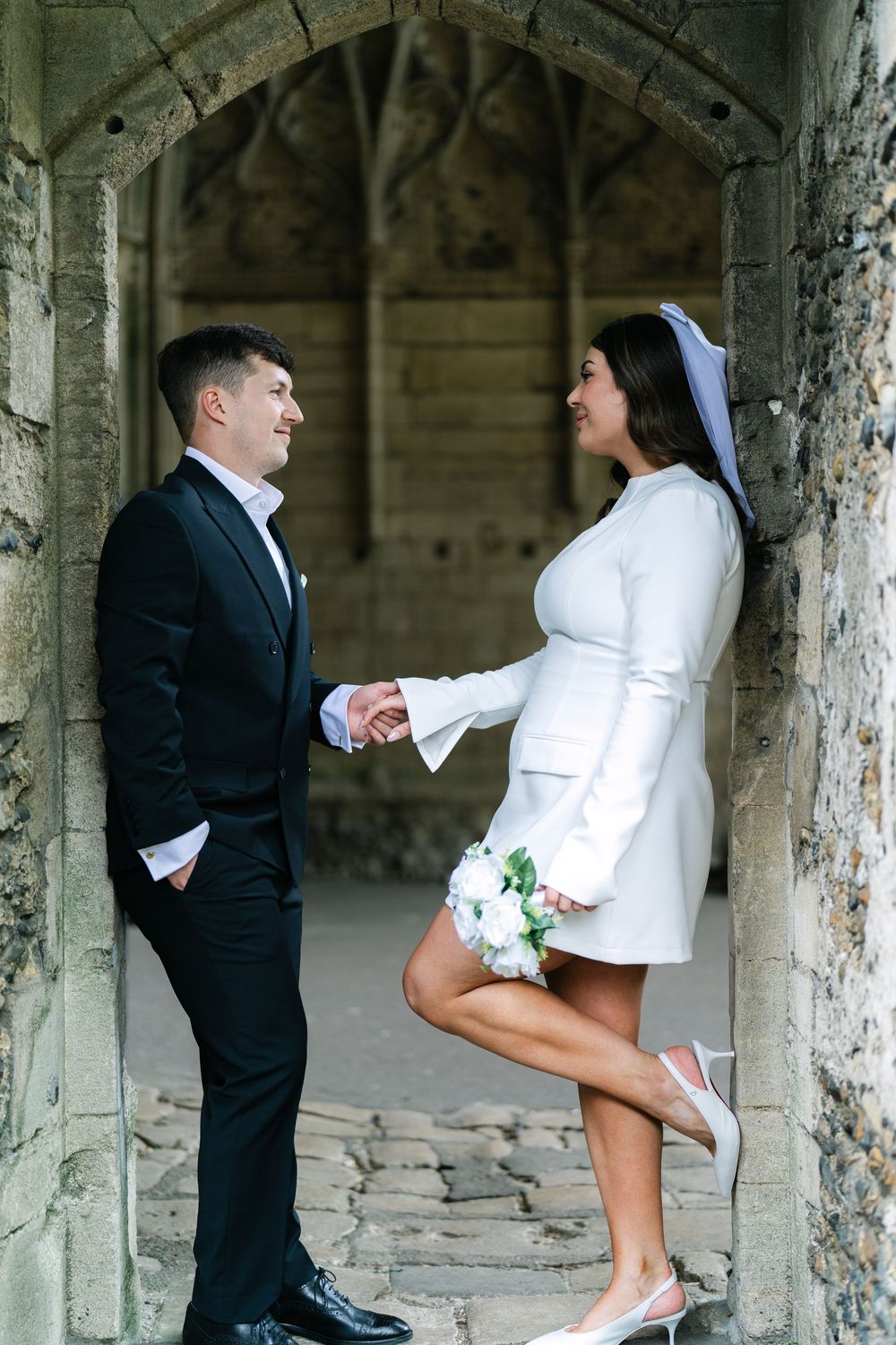 Wedding party poses playfully in a stone colonnade with bride wearing white coat and groom in black suit.