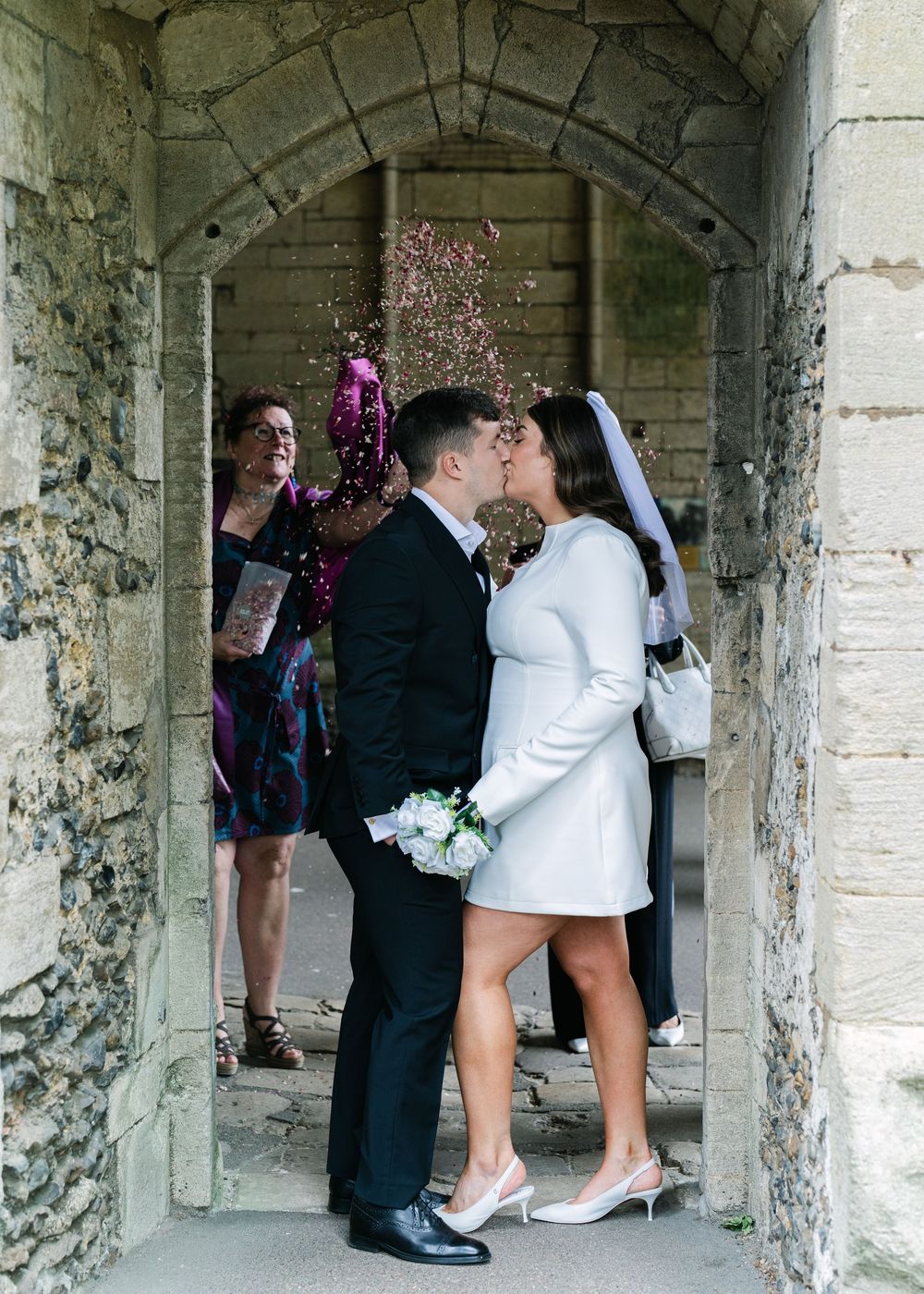 A couple shares a kiss in a stone archway while someone throws confetti during their winter wedding celebration.