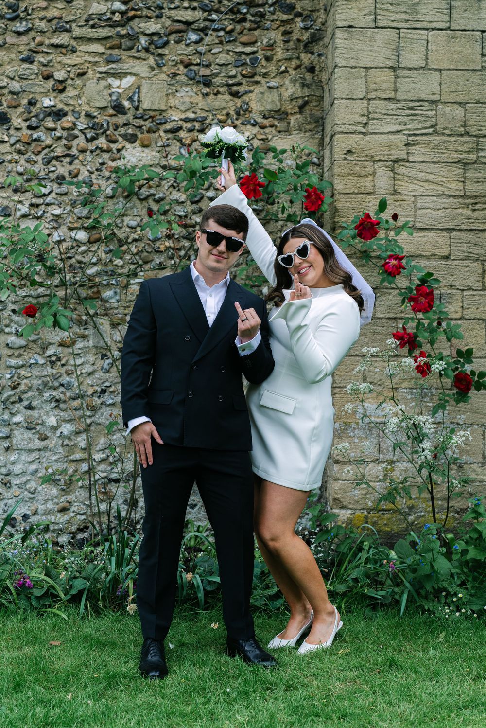 A stylishly dressed couple poses playfully against a stone wall with climbing red roses in an outdoor garden setting.