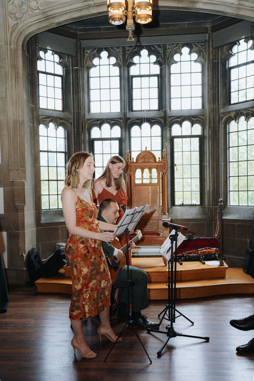 Musicians perform in an elegant Gothic room with tall arched windows.